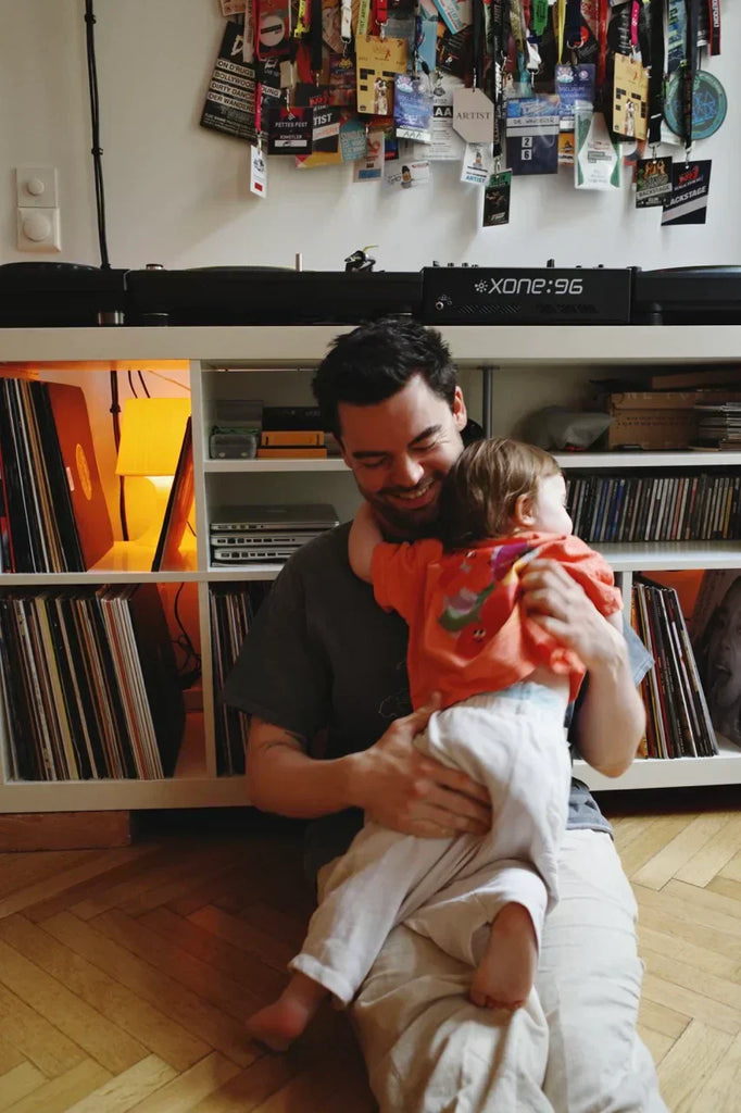 DJ Max Wanderer sitting on the floor at home, smiling as he holds his young daughter Emma in front of a vinyl record collection and DJ setup.