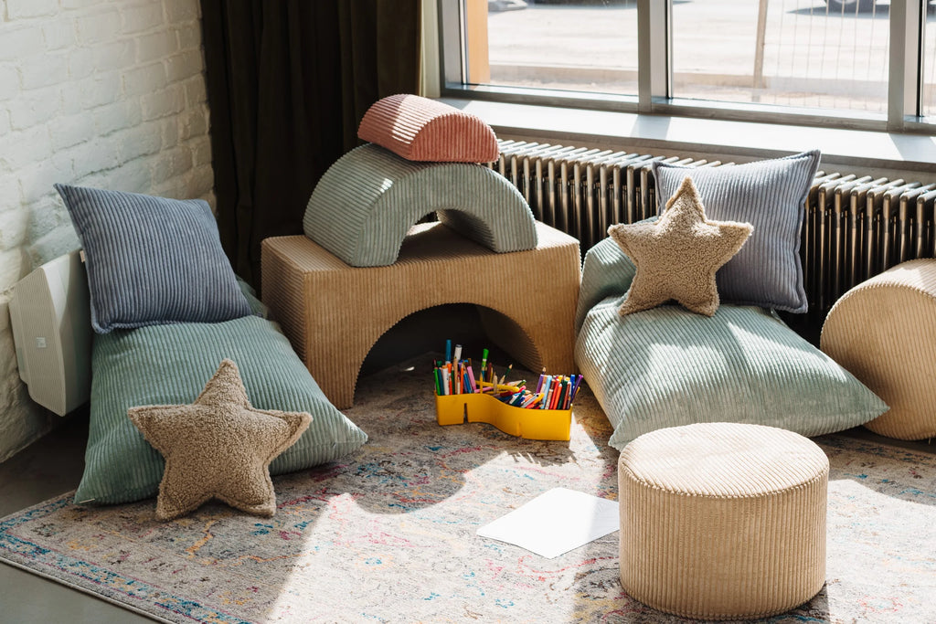 A cosy corner setup featuring Wigiwama’s corduroy play furniture and pillows in muted tones, arranged near a sunny window with art supplies and a playful star cushion on the floor.