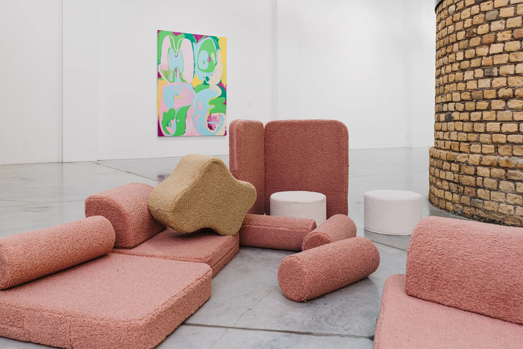 Wigiwama soft play blocks in blush pink and sand tones arranged in a modern gallery space, placed in front of a colourful “HOPE” painting by Erwin Wrum on a white wall.