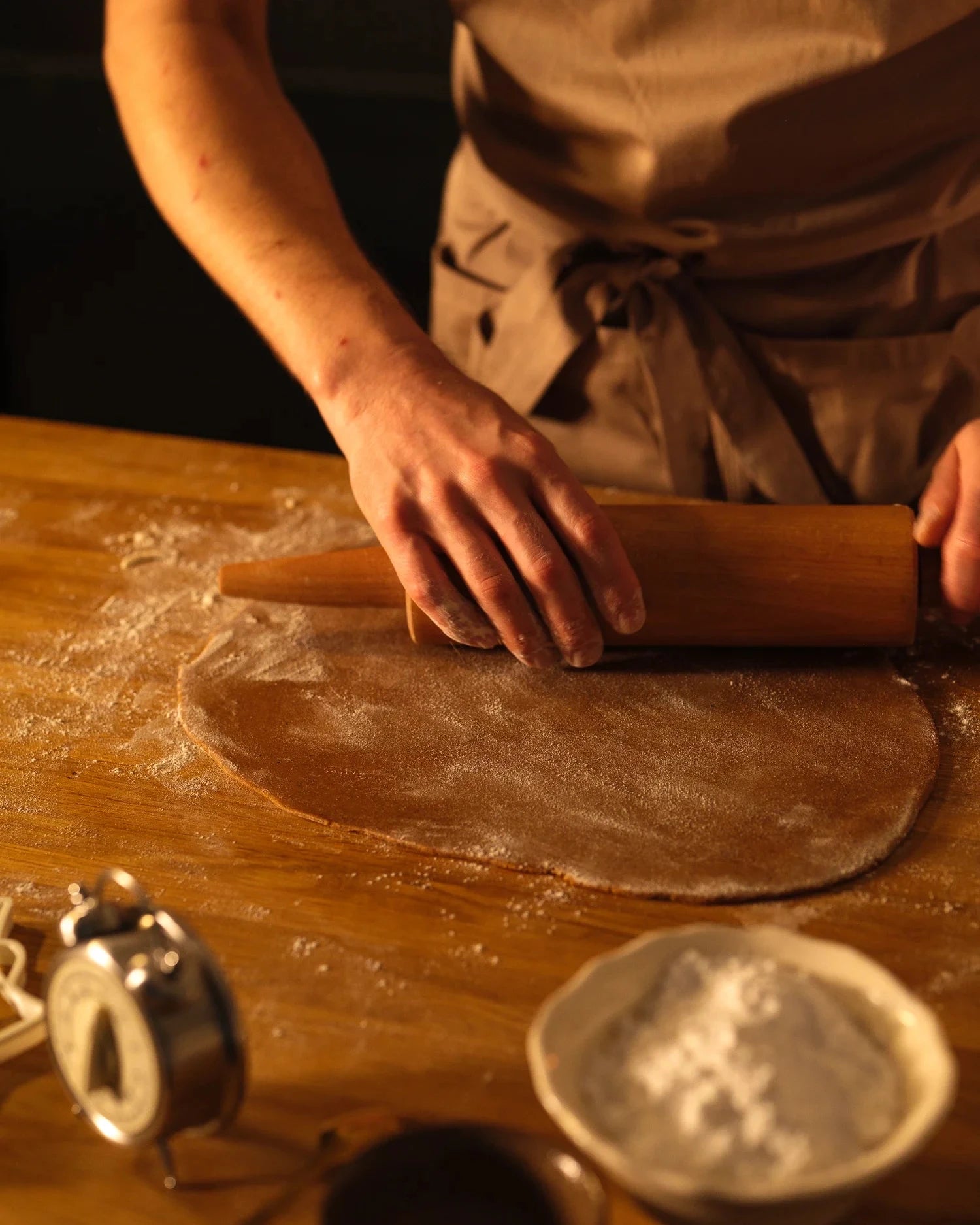 Person rolling out dough on a wooden surface with a bowl of flour and a timer in the background.