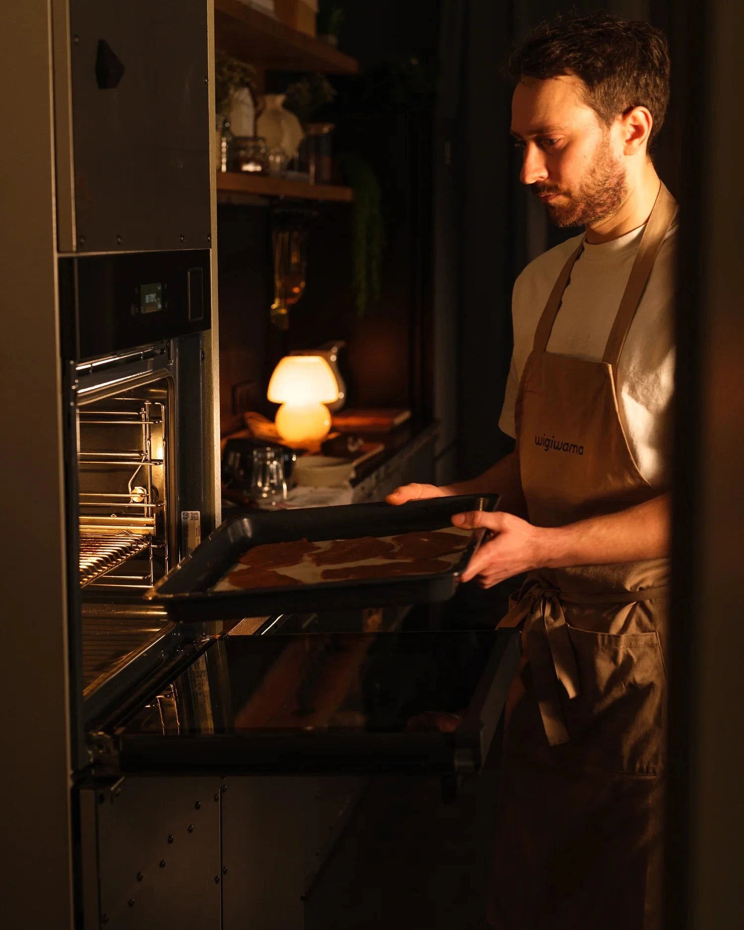 Person wearing an apron taking a tray out of an oven in a kitchen setting.