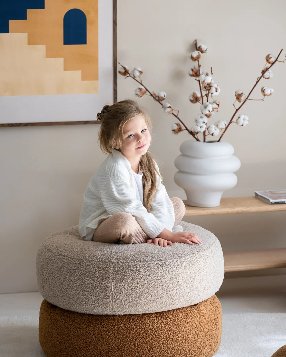 Young girl sitting on a round ottoman in a room with a vase of cotton branches and abstract art.