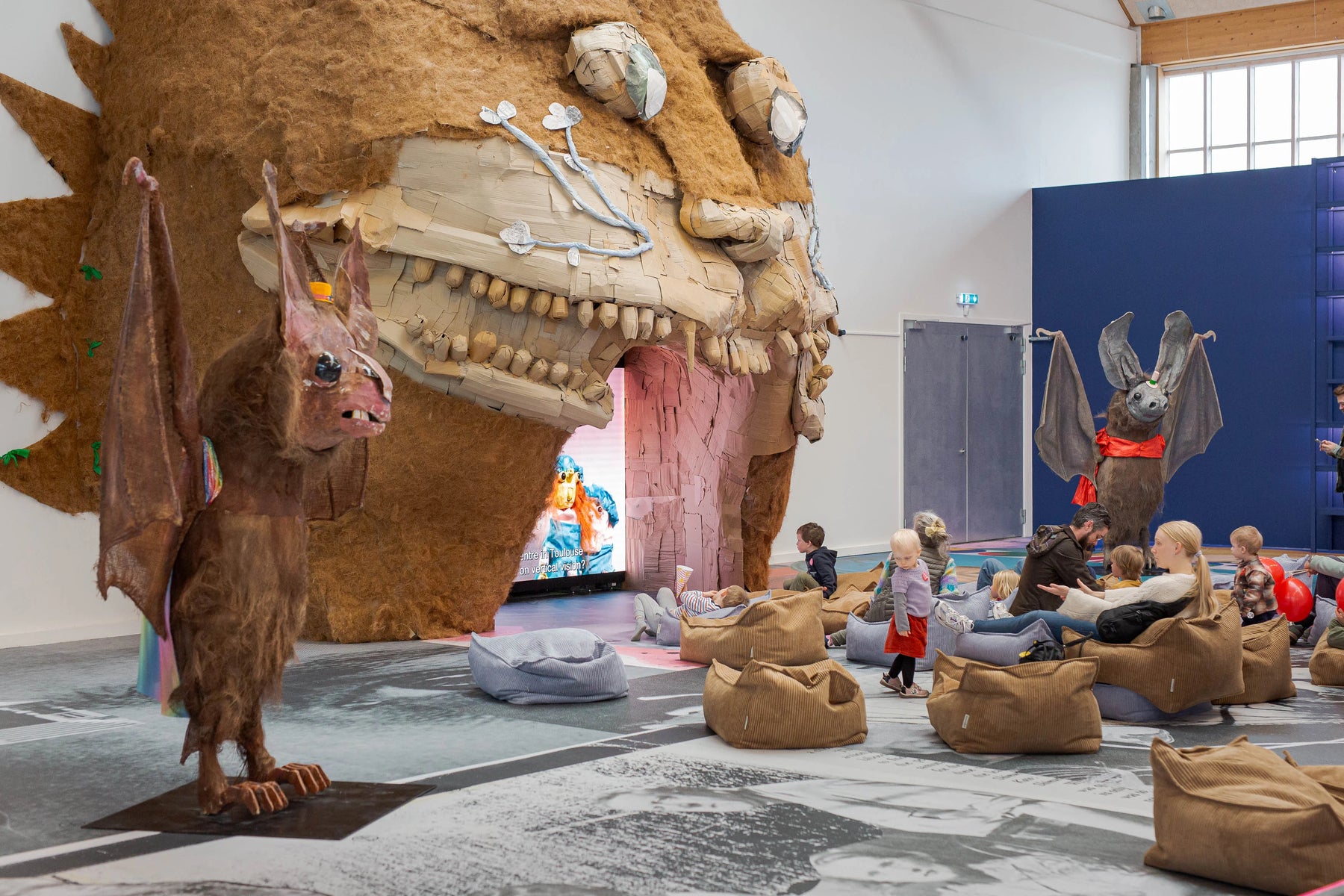 Children and adults sitting on Wigiwama beanbags surrounded by bat sculptures inside the immersive exhibition by Monster Chetwynd at Copenhagen Contemporary