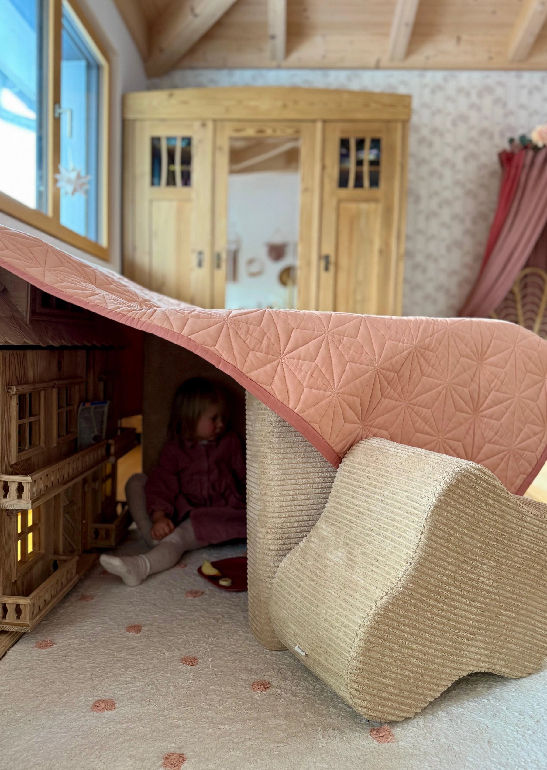 Child hiding under a play fort made from Wigiwama soft play pieces and a quilted throw in a wooden kids room
