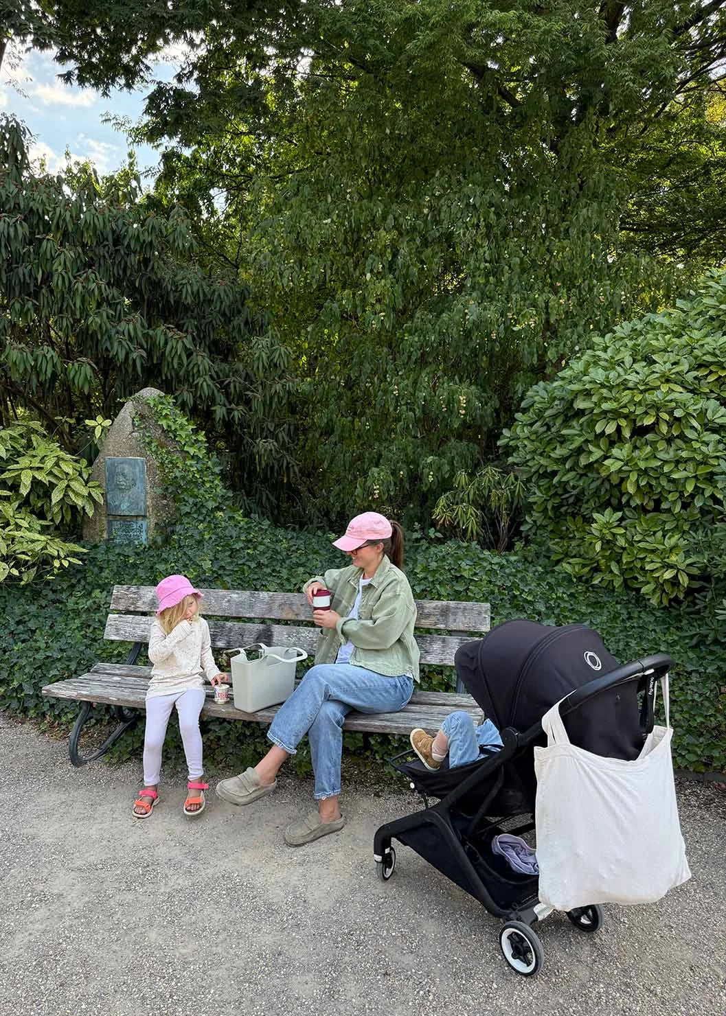 A mother and daughter sit on a bench with snacks while a stroller stands nearby in the green surroundings of the Botanical Garden.