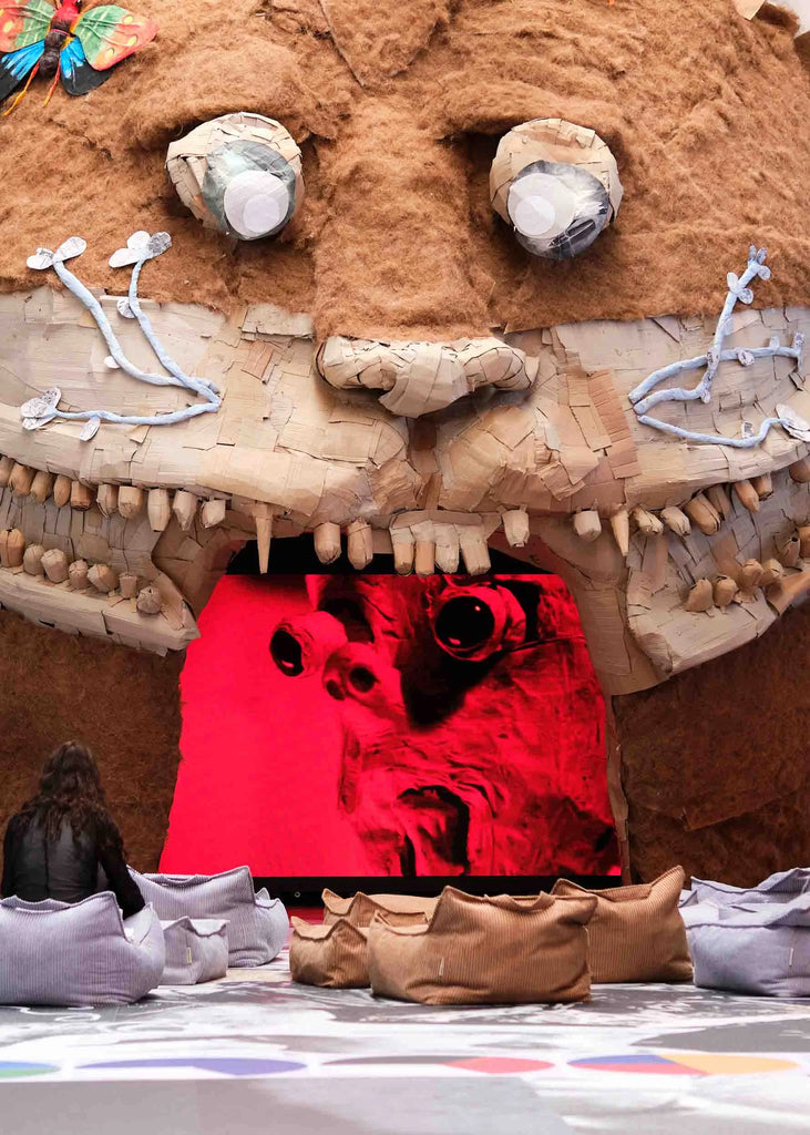 A woman sits on Wigiwama corduroy beanbags in front of a large monster-like sculpture made from natural materials, watching a surreal red-toned video playing inside its mouth at Monster Chetwynd’s exhibition at Copenhagen Contemporary.