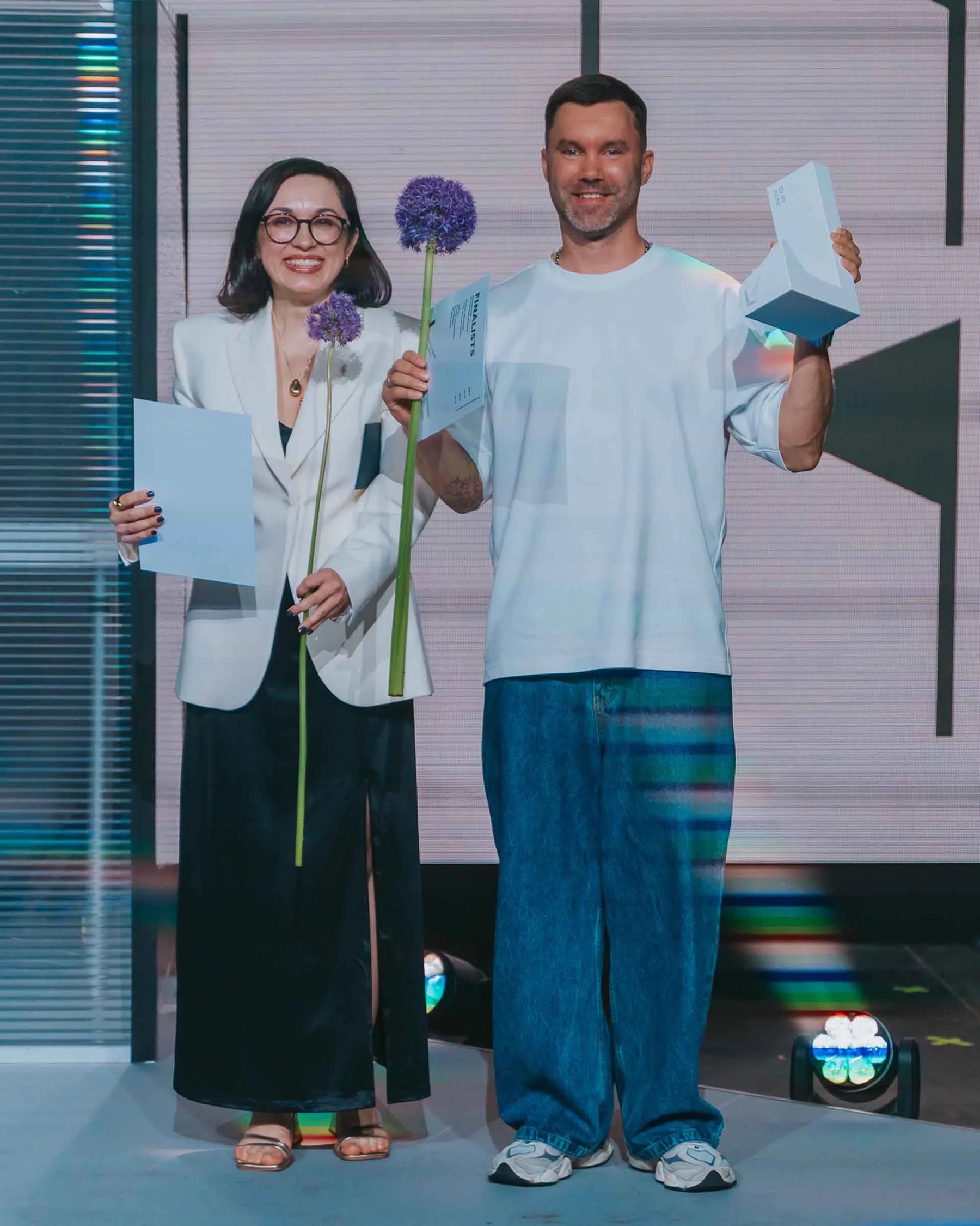 Mark Litvyakov and Baiba Lindane on stage holding the National Design Award of Latvia 2025 trophies and flowers after winning in the Product Design category.