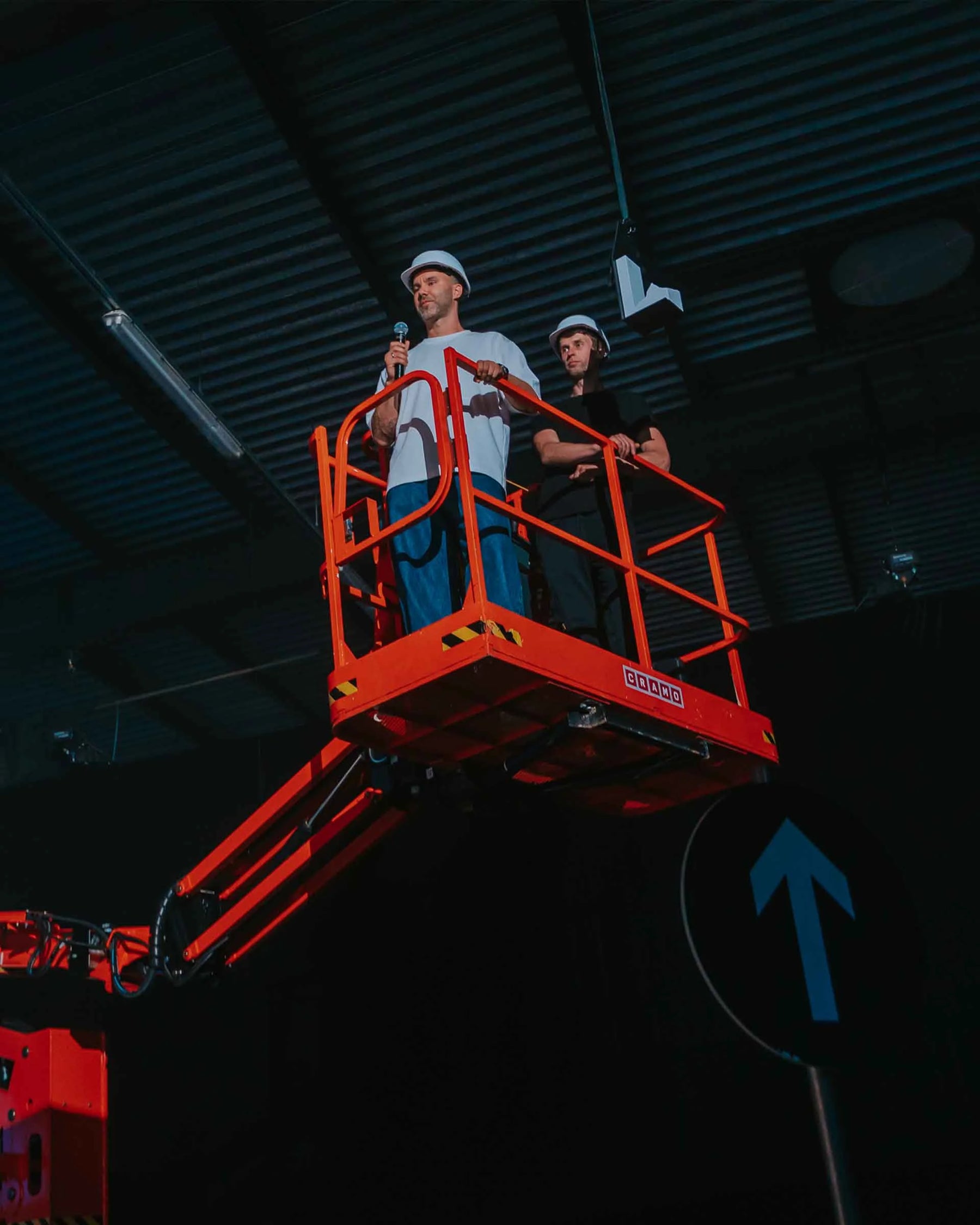 Product designer Mark Litvyakov speaking and receiving the award at the National Design Award of Latvia 2025 ceremony, elevated on a scissor lift in an industrial-style venue.