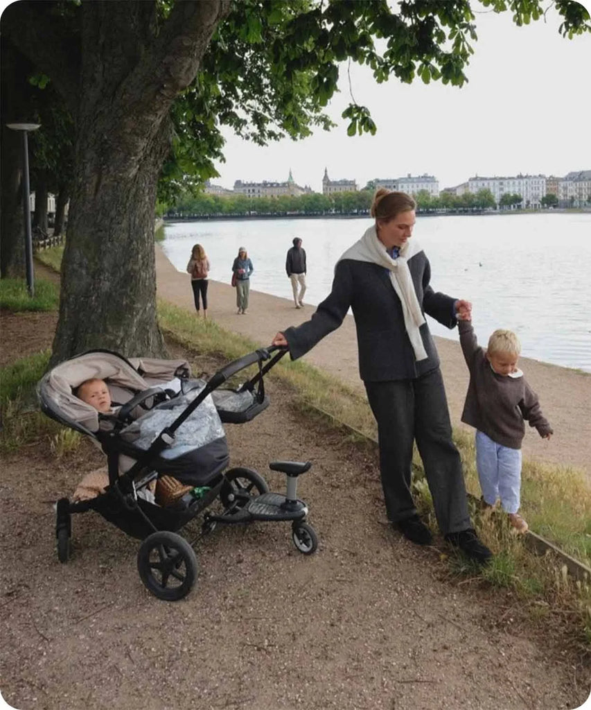 Lea Sørensen walks along a lakeside path in Copenhagen, holding her son's hand and pushing a stroller under a large tree.