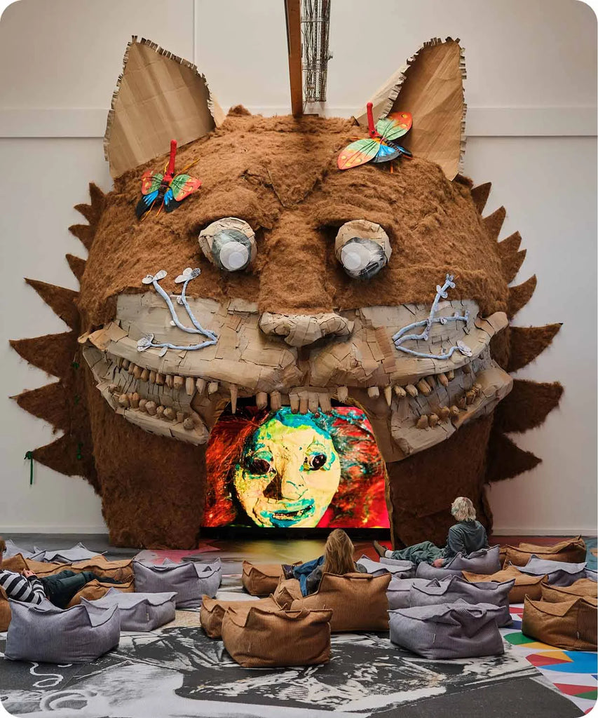 Visitors sit on beanbags in front of a large, whimsical monster-shaped installation with a screen inside its mouth, displaying colourful animated art at Copenhagen Contemporary.