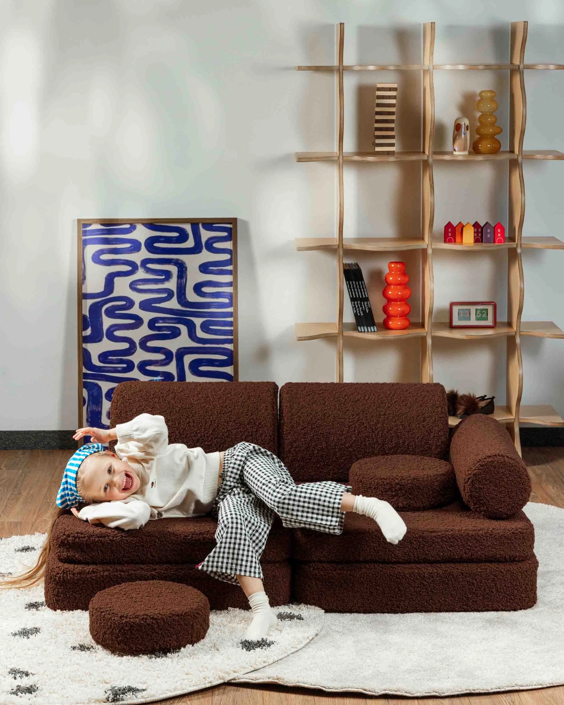 Child lying on a brown sofa in a room with a bookshelf and abstract wall art.