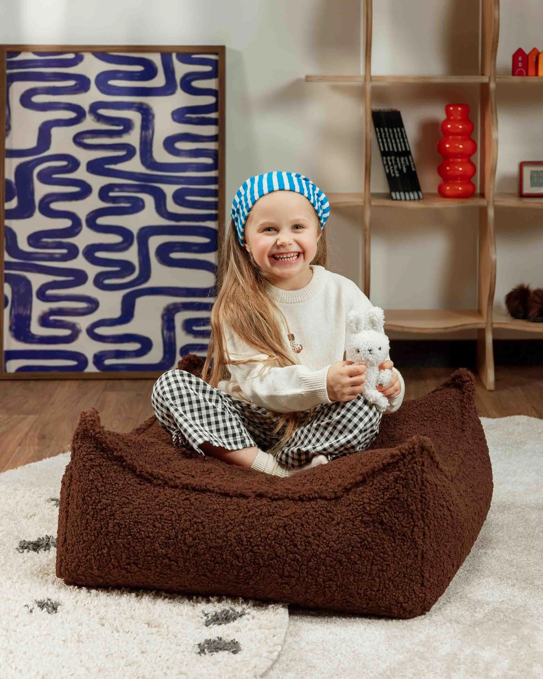 Child sitting on a brown ottoman holding a white teddy bear, with a patterned wall and shelves in the background.