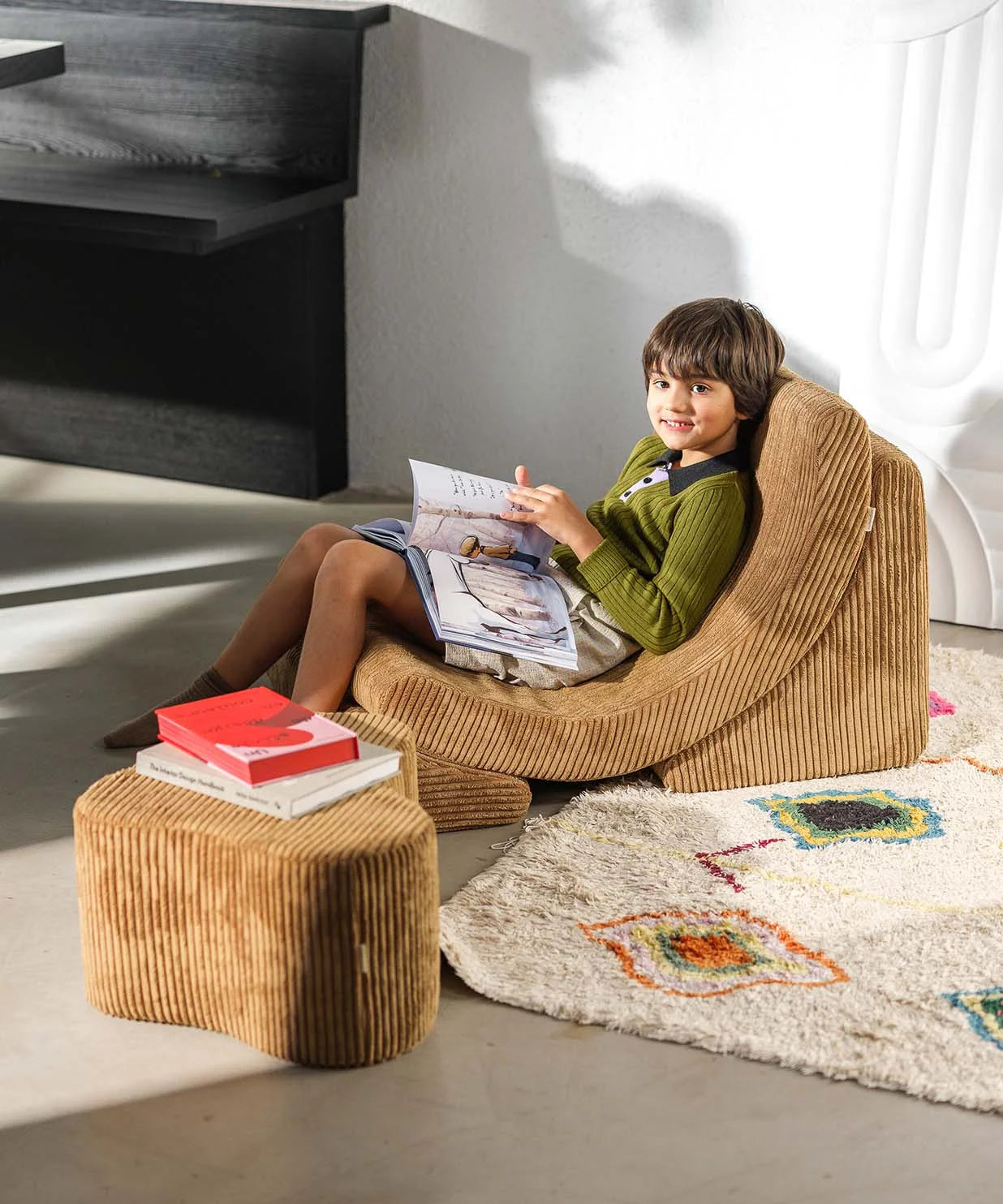Child sitting on a brown ribbed chair reading a magazine in a modern living room.