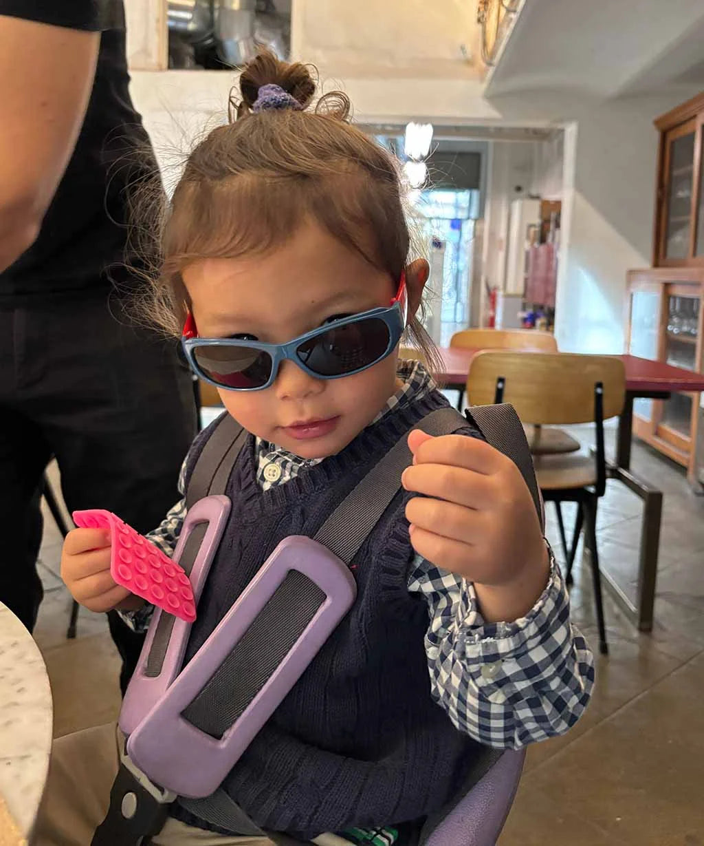 Child wearing sunglasses and a purple seatbelt in a casual indoor setting